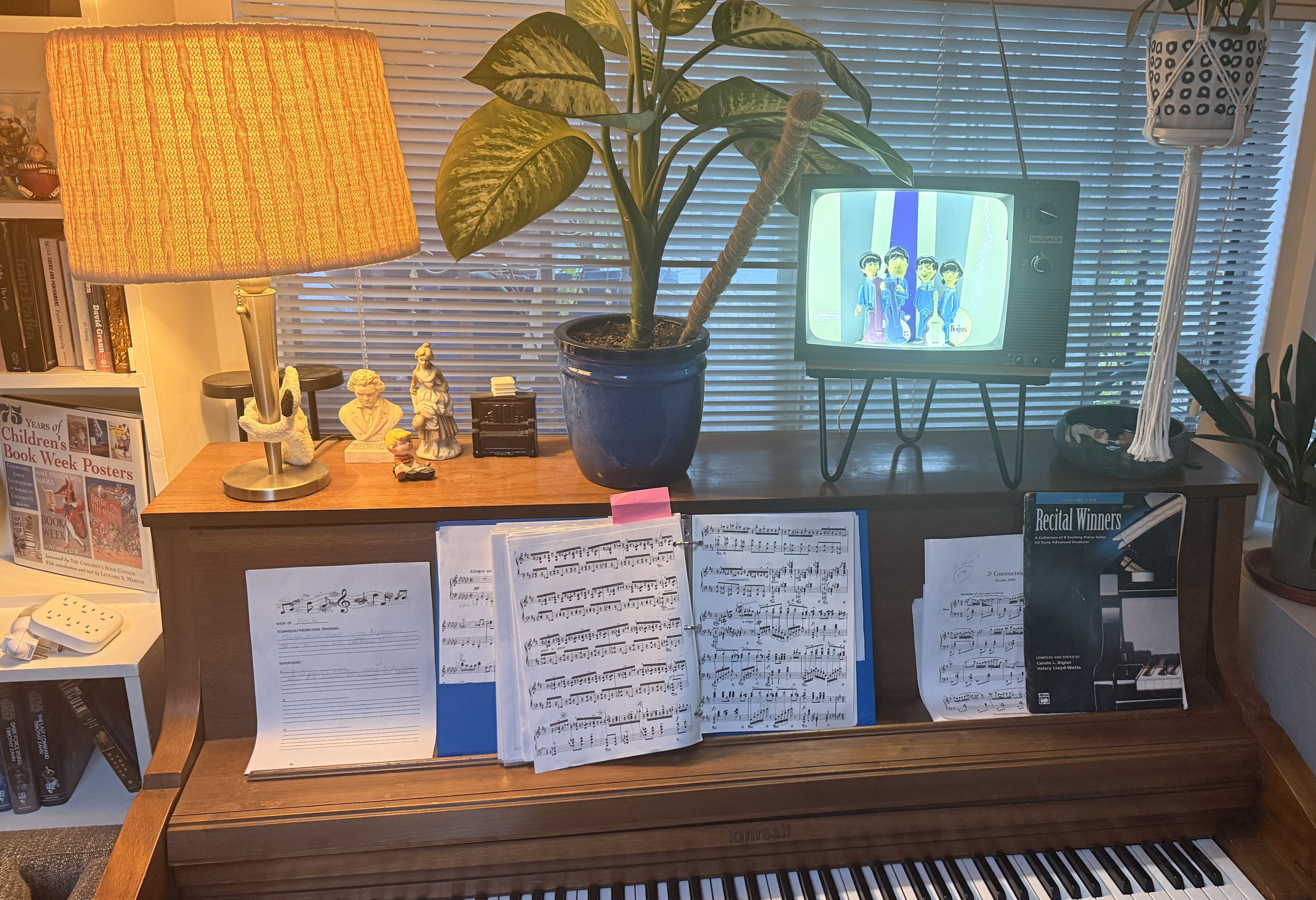 a small and illuminated retro looking tv set with figurines of the Beatles and a  striped gray and blue background sits on top of a piano next to a plant the room has a 70's vibe